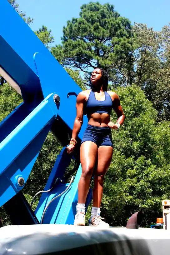 A muscular woman in a blue sports bra and shorts stands confidently atop a large blue construction lift arm outdoors…
