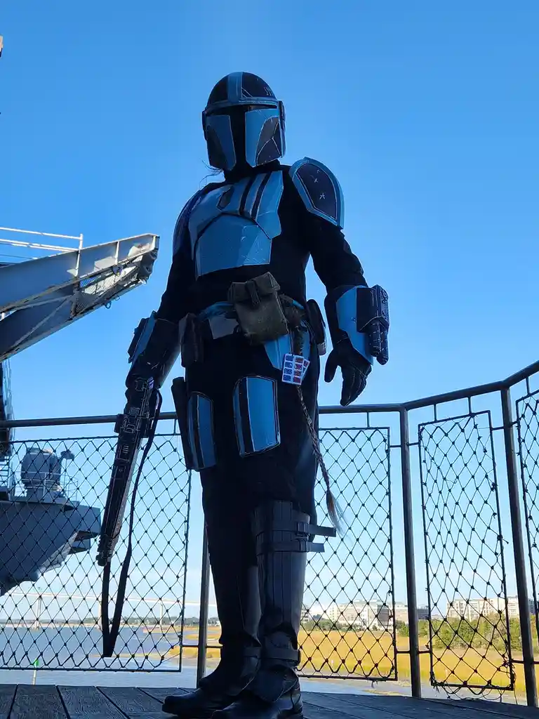 A person in a detailed blue and black Mandalorian armor costume stands on a wooden deck behind a chain-link fence, holding a…
