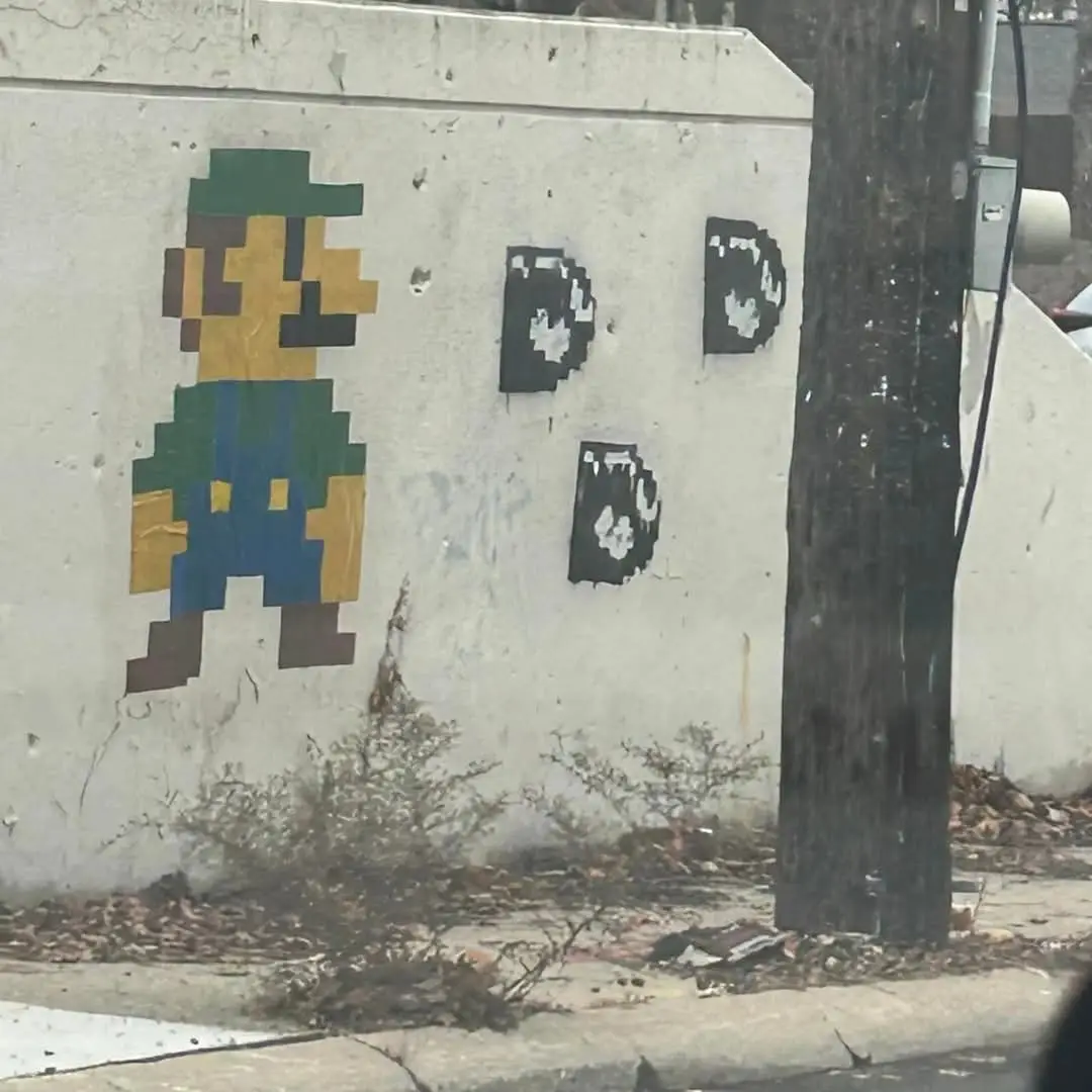 A pixelated mural of the video game character Mario is painted on a weathered concrete wall next to a utility pole. To the…