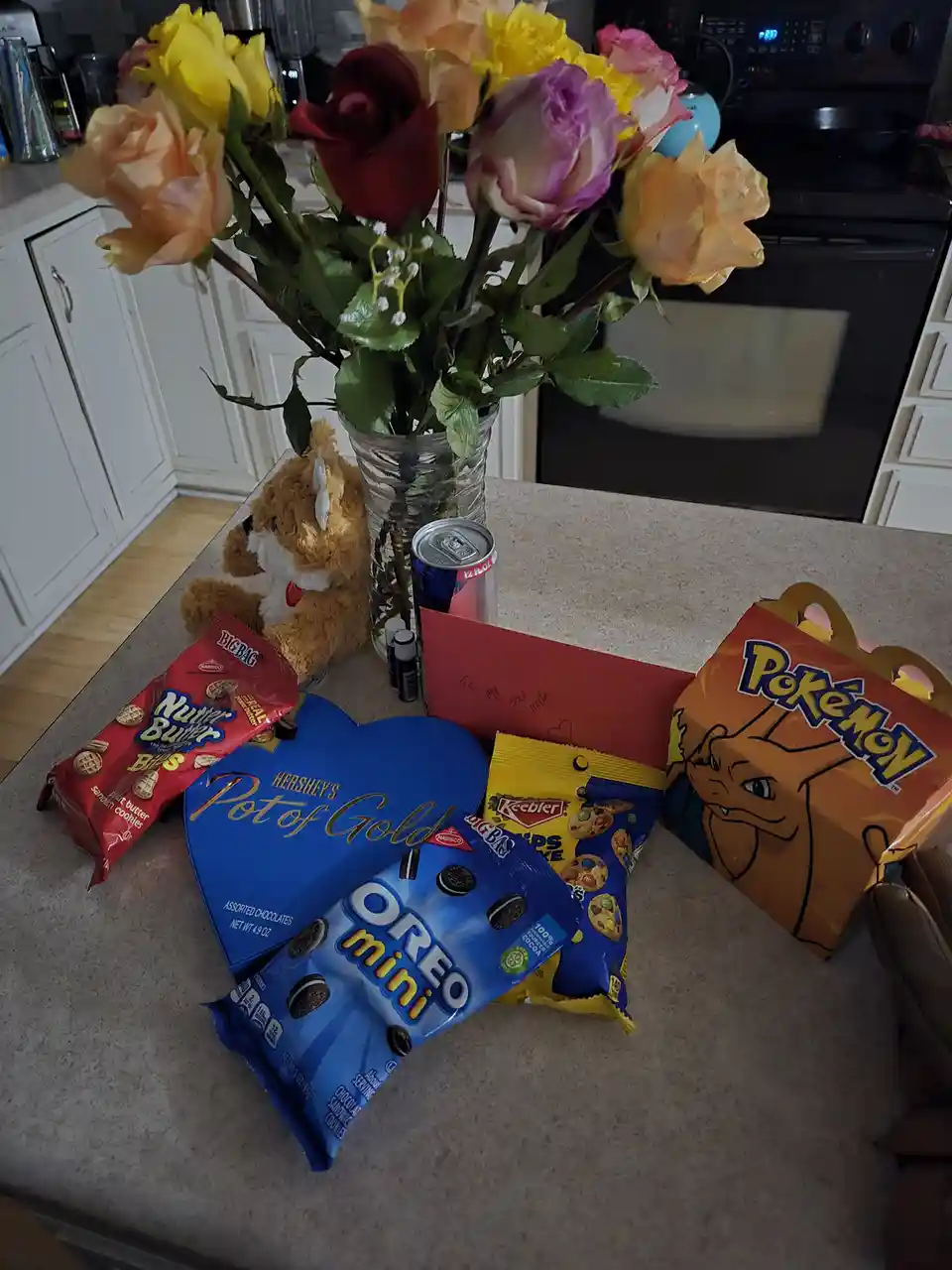A kitchen counter displays a bouquet of colorful roses in a glass vase, surrounded by various snacks including a…