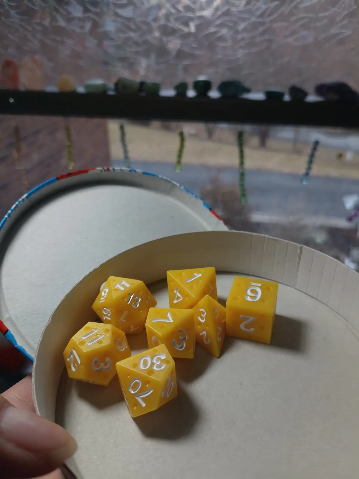 A hand holds a paper cup containing several yellow polyhedral dice with silver numbers, showing results like 1, 2, 3, 4, 6…