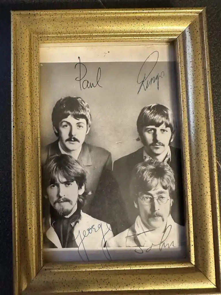 A black-and-white photograph of the Beatles, framed in gold, with handwritten signatures above each member's head: 'Paul'…