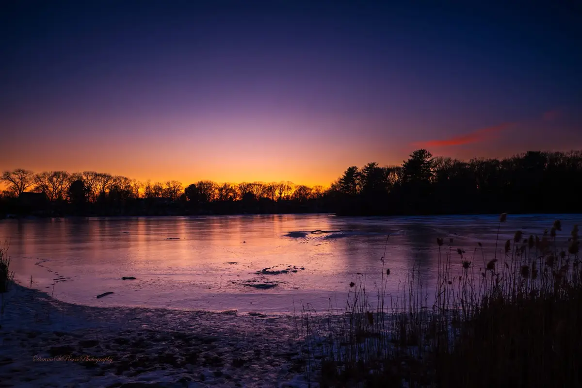 A serene winter sunset over a frozen lake, with vibrant orange and purple hues reflecting on the ice. Silhouetted trees line…