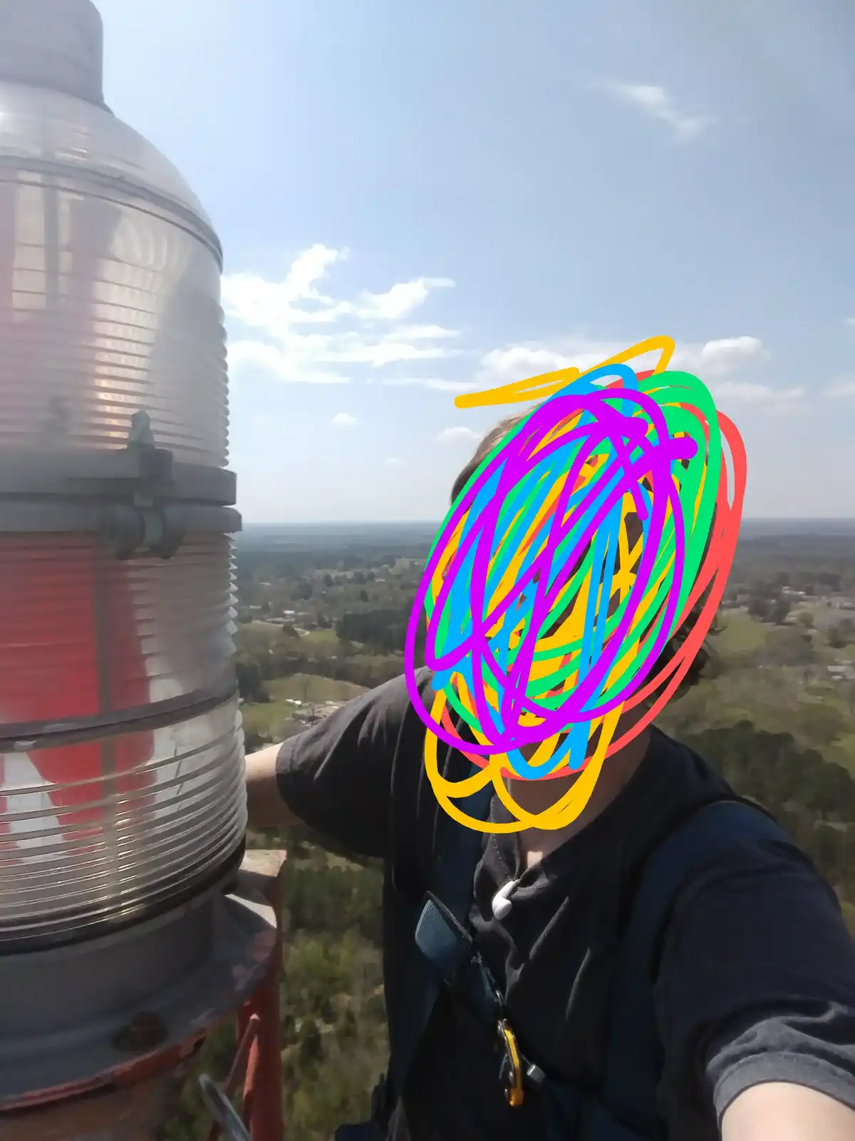 A person wearing a black shirt and safety harness stands atop a lighthouse, taking a selfie with a large, ribbed lighthouse…