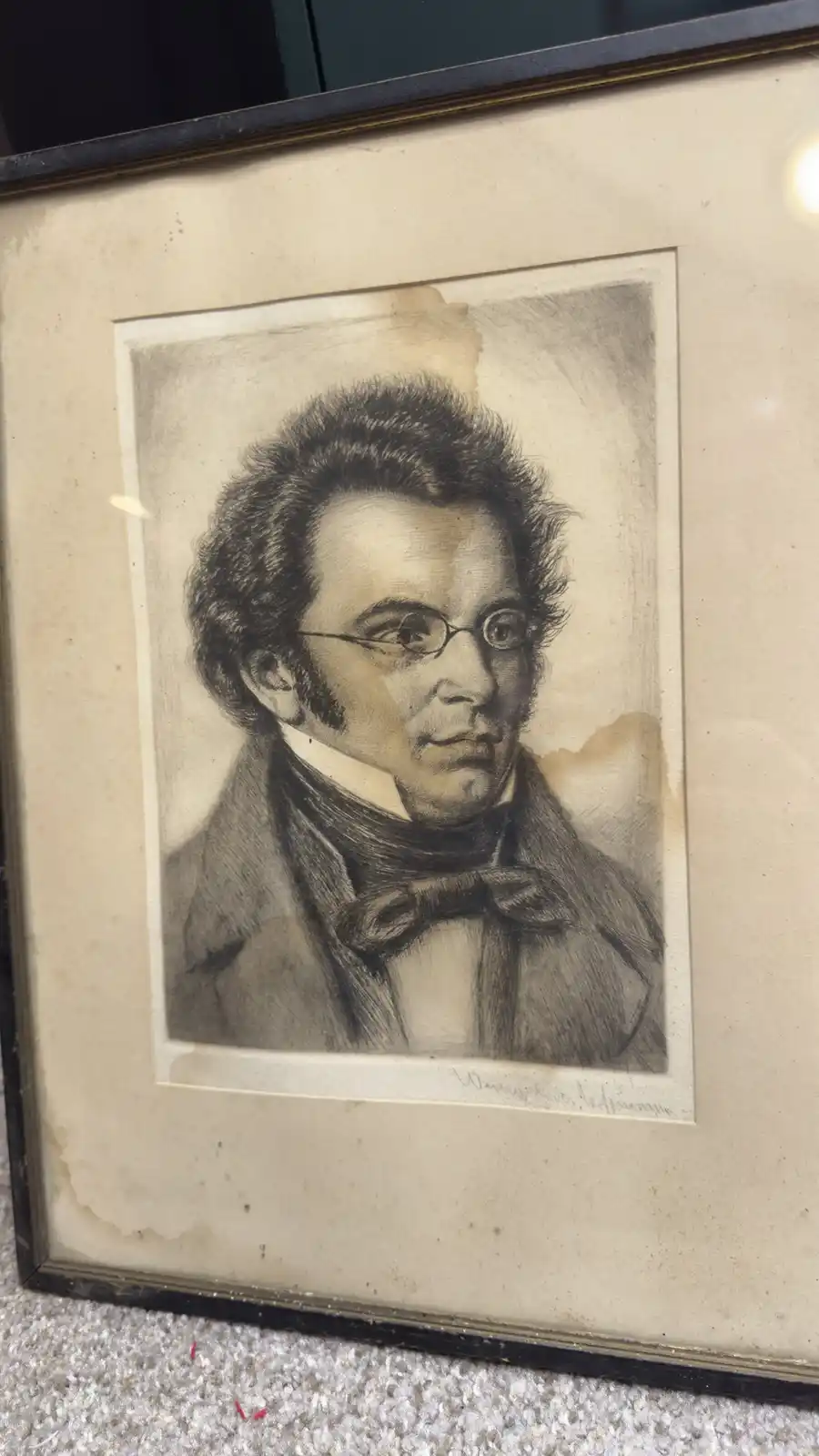 A framed, aged charcoal portrait of a man with curly hair, glasses, and a bow tie, showing visible water damage on the…