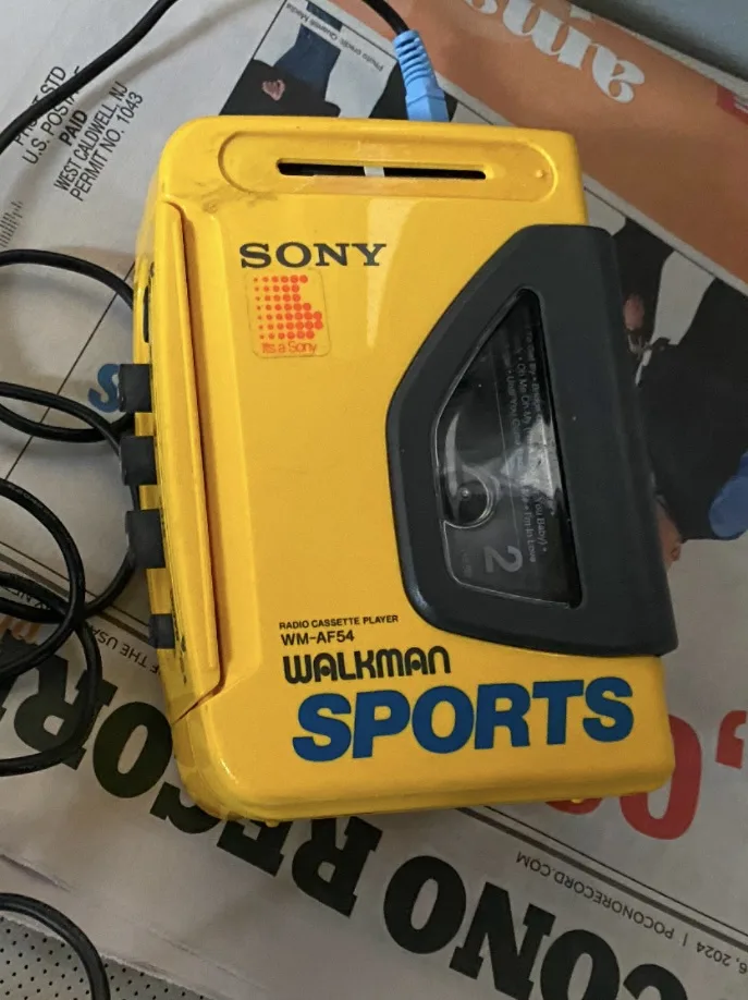 A yellow Sony Walkman Sports WM-AF54 radio cassette player with a visible cassette tape inside, resting on a newspaper. The…