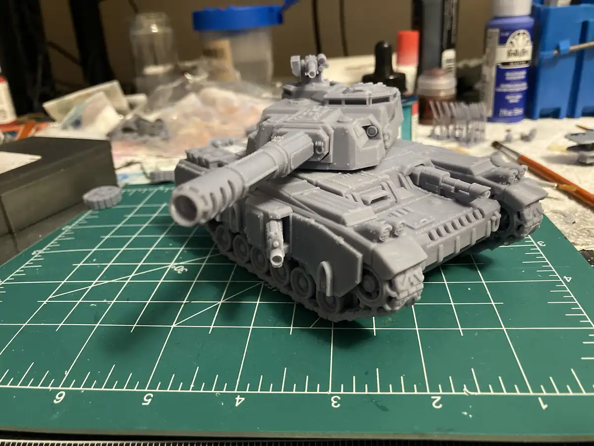 A gray, unpainted plastic model tank sits on a green cutting mat with grid lines and measurements, surrounded by hobby…