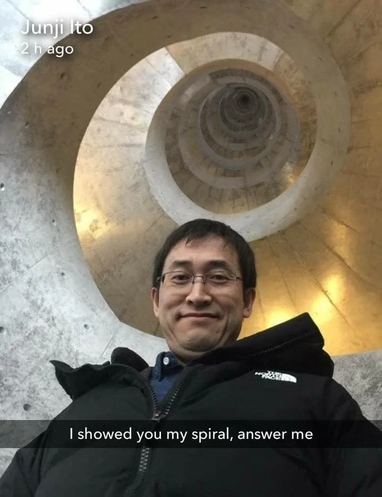 A man with glasses and a black jacket takes a selfie in front of a large, spiraling concrete architectural structure. The…