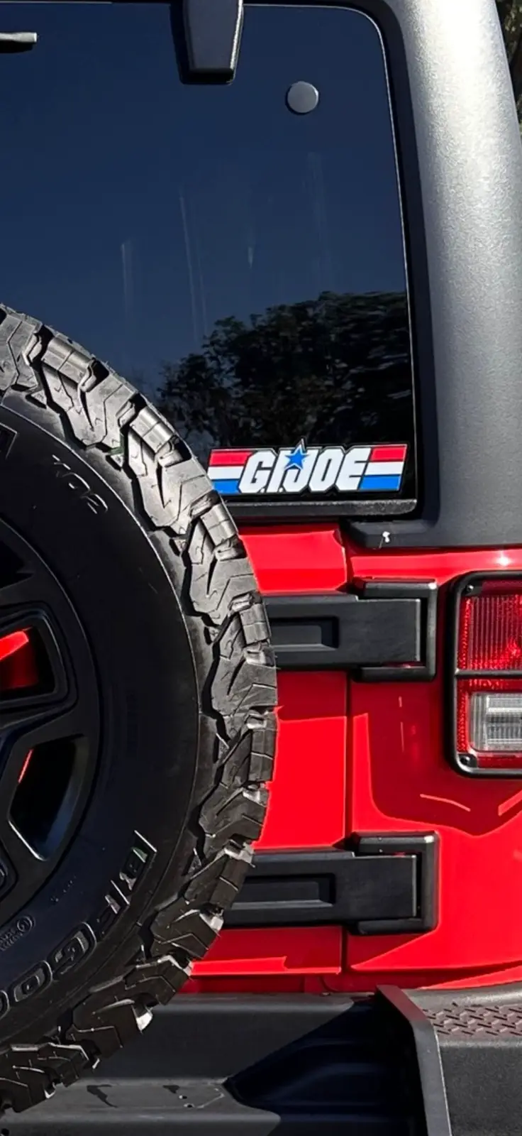 A close-up of the rear of a red Jeep, showing a spare tire mounted on the back and a 'G.I. JOE' sticker affixed to the rear…