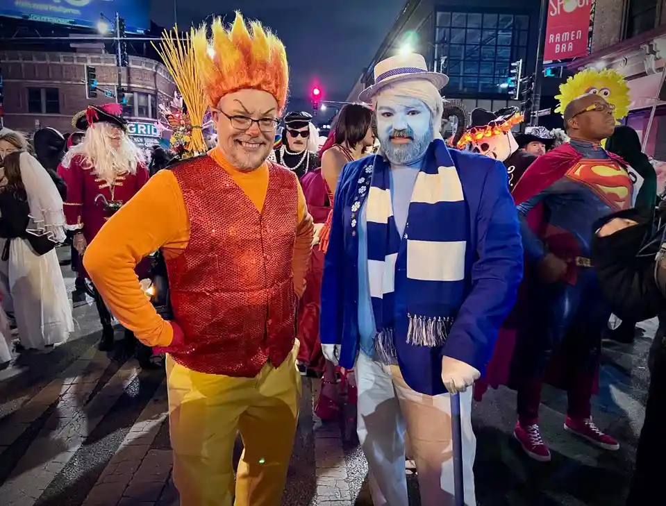 Two men in elaborate costumes stand side-by-side at a nighttime street event. The man on the left wears a fiery orange wig…
