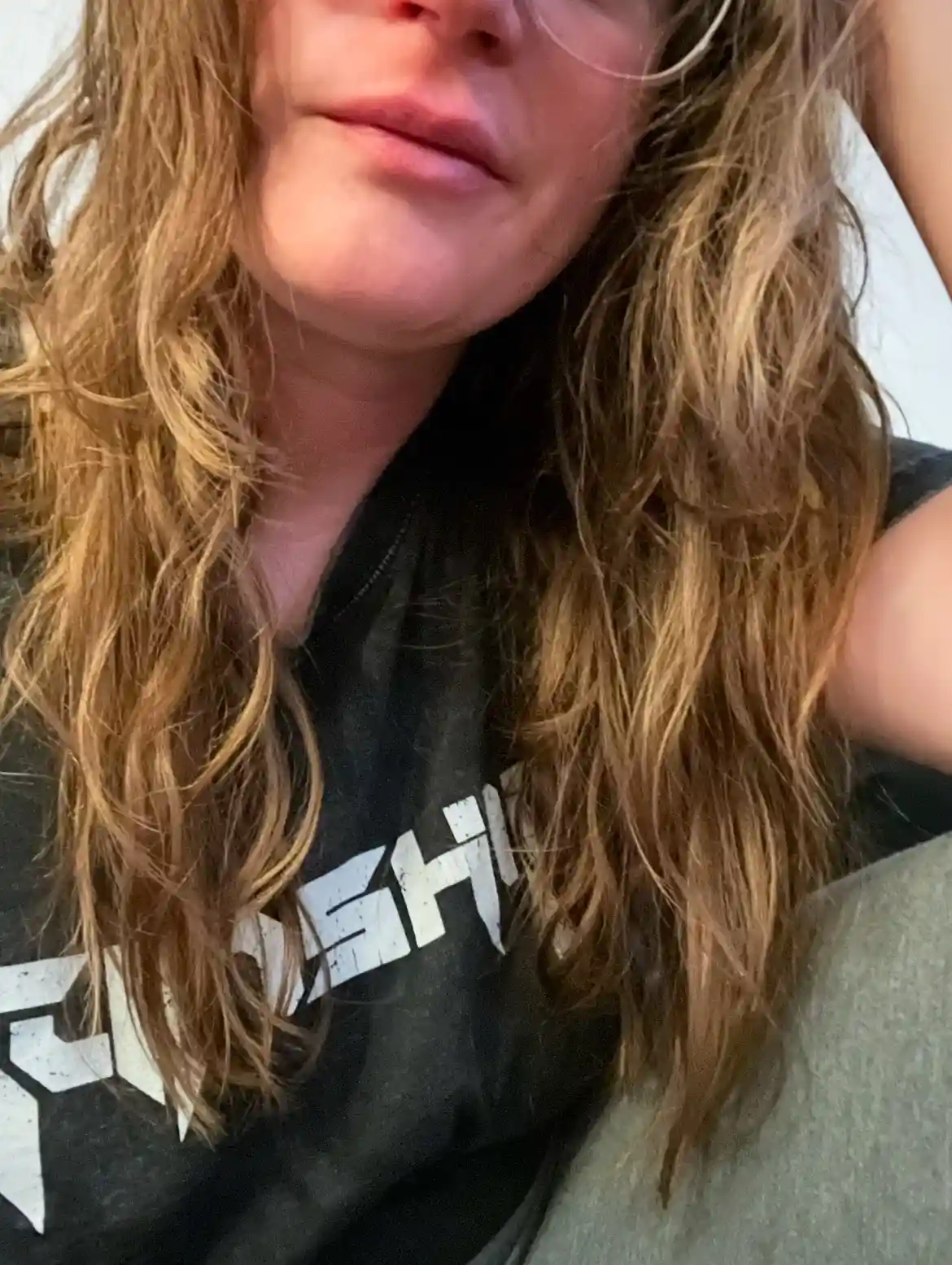 A close-up selfie of a person with long, wavy, light brown hair, wearing a black t-shirt with white text partially obscured…