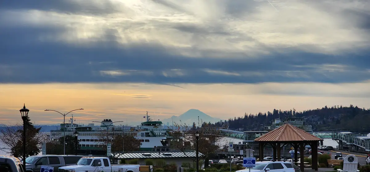 A scenic view of a waterfront ferry terminal at dusk, with a large ferry docked, a gazebo in the foreground, and a distant…