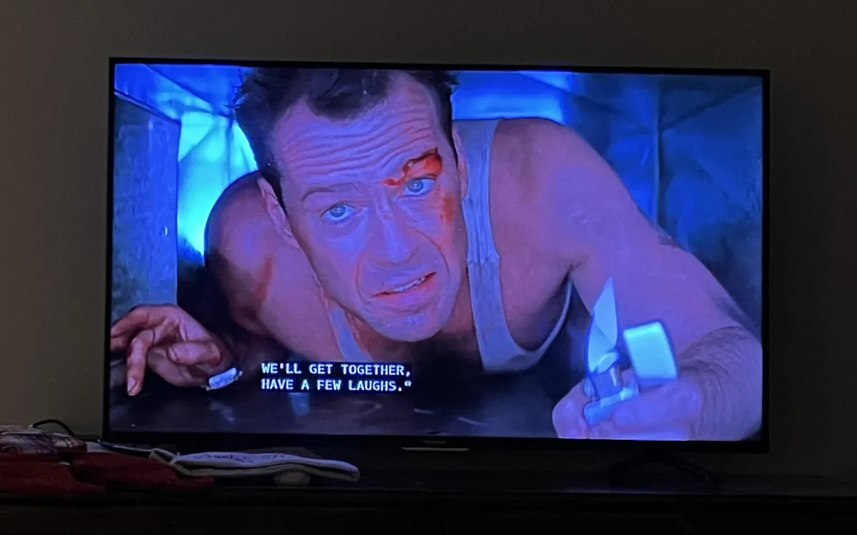 A television screen shows a close-up of a man in a white tank top with a bloody eye and face, holding a knife and a lighter…