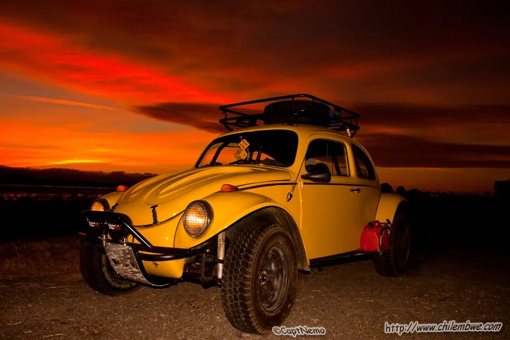 A yellow, modified Volkswagen Beetle with oversized tires, a roof rack, and a red gas can is parked on a dirt road under a…
