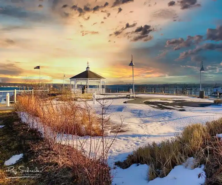 A scenic winter photo shows a snow-covered gazebo and walkway at sunset, with an American flag and distant water visible…