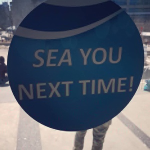 A blue circular sign with white text reading 'SEA YOU NEXT TIME!' is held up, likely by a person in camouflage pants, with a…