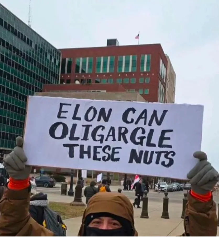 A person wearing a brown hooded jacket and gloves holds up a handmade white sign with black marker text reading 'ELON CAN…