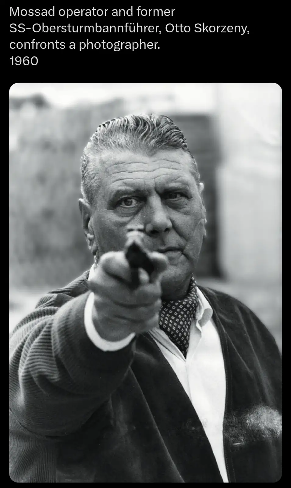 A black-and-white photograph shows Otto Skorzeny, a man with slicked-back hair and a stern expression, pointing a handgun…