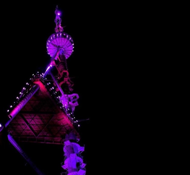 A nighttime photo of a tall, illuminated tower structure, possibly a communications or observation tower, bathed in purple…