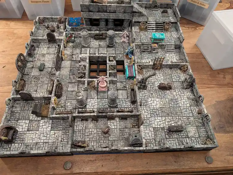 An overhead view of a detailed, handcrafted miniature dungeon map made of grey stone tiles with various obstacles, doors…