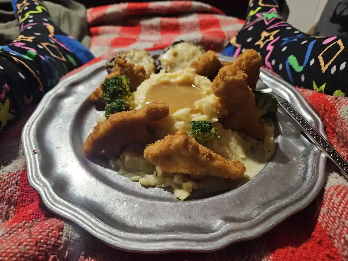 A close-up photo shows a silver plate with mashed potatoes, gravy, fried chicken tenders, and broccoli, resting on a red…