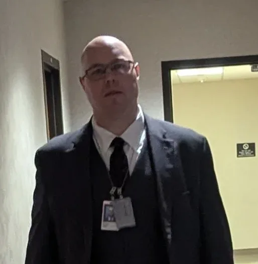 A bald man in glasses, a dark suit, white shirt, and tie stands in a hallway wearing a security badge. He has a neutral…