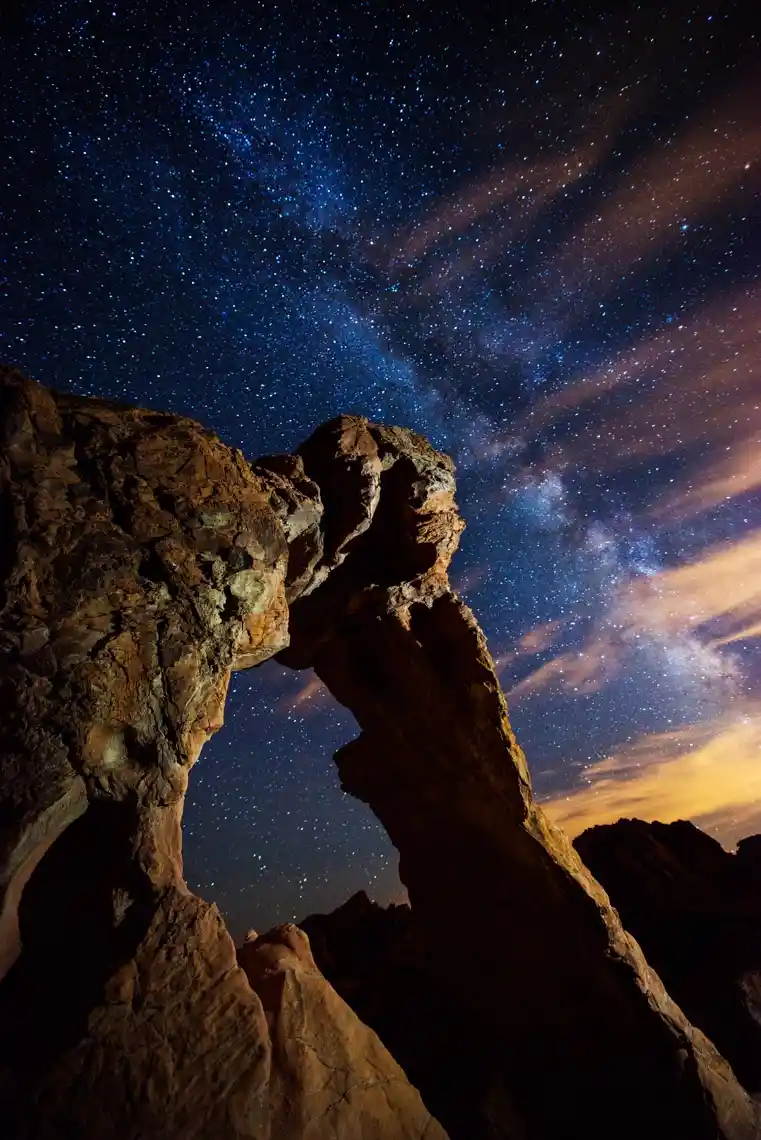 A low-angle night photograph shows a rugged, natural rock arch silhouetted against a vibrant, star-filled sky with visible…