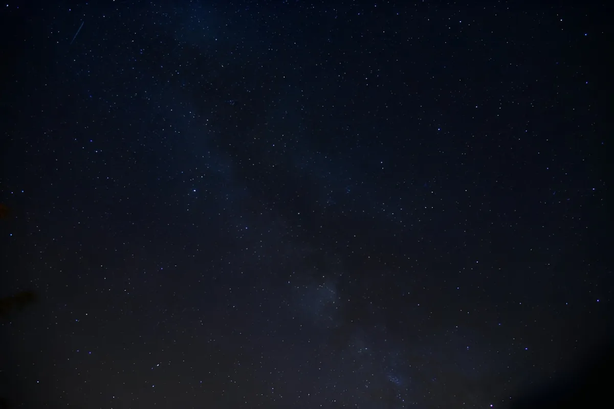 A dark night sky filled with countless stars and the faint, glowing band of the Milky Way galaxy. The image has no people or…