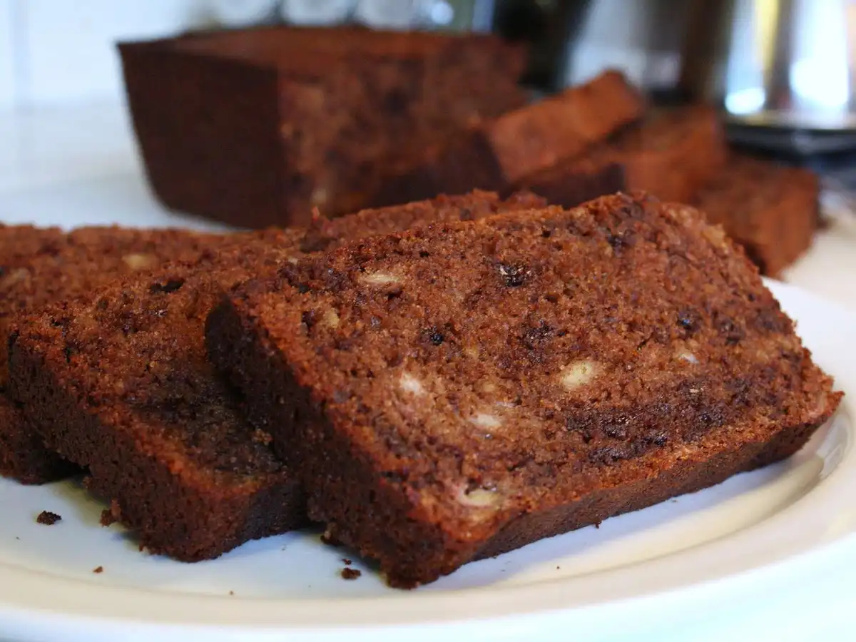 A close-up photo shows several thick slices of dark brown banana bread on a white plate, with the rest of the loaf visible…