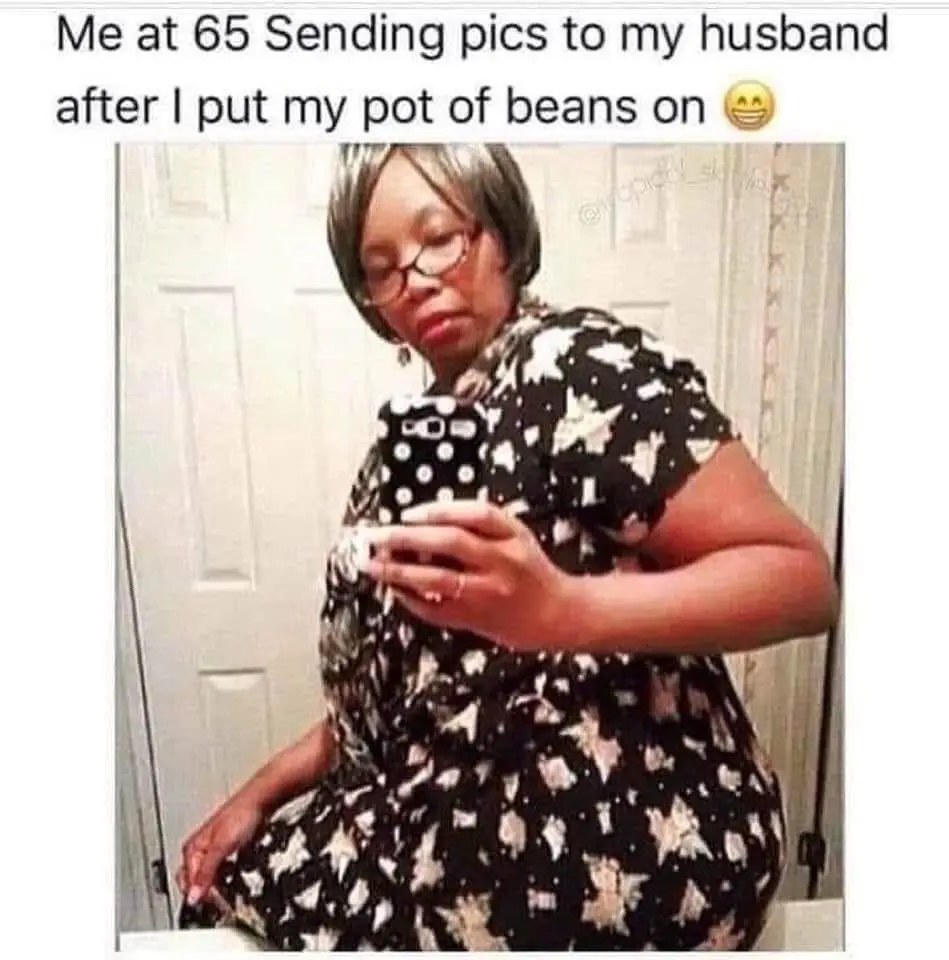 A woman with short gray hair and glasses takes a selfie in a mirror while holding a smartphone with a polka-dot case. She…