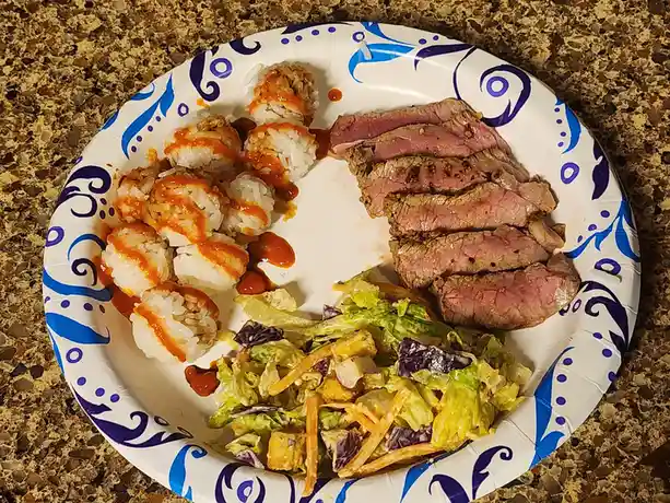 A paper plate with blue swirl patterns holds a meal of sliced medium-rare steak, a side salad with shredded carrots and…