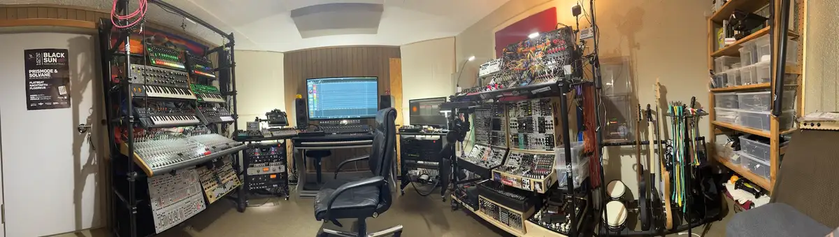 A panoramic view of a cluttered, high-tech music studio filled with racks of analog synthesizers, modular gear, mixing…