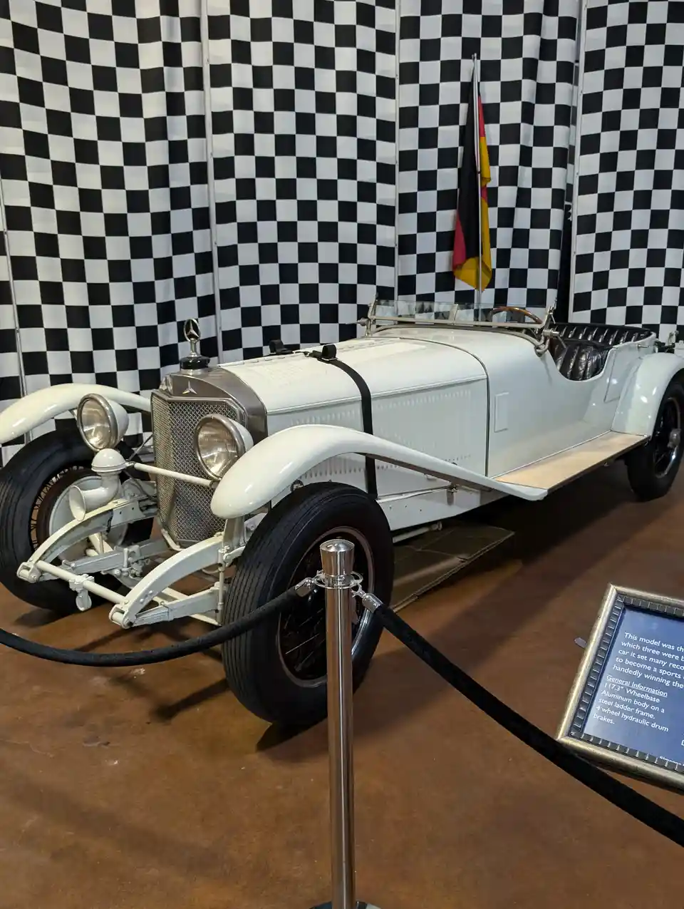 A white vintage Mercedes-Benz race car is displayed in a museum with a black-and-white checkered backdrop and a German flag…