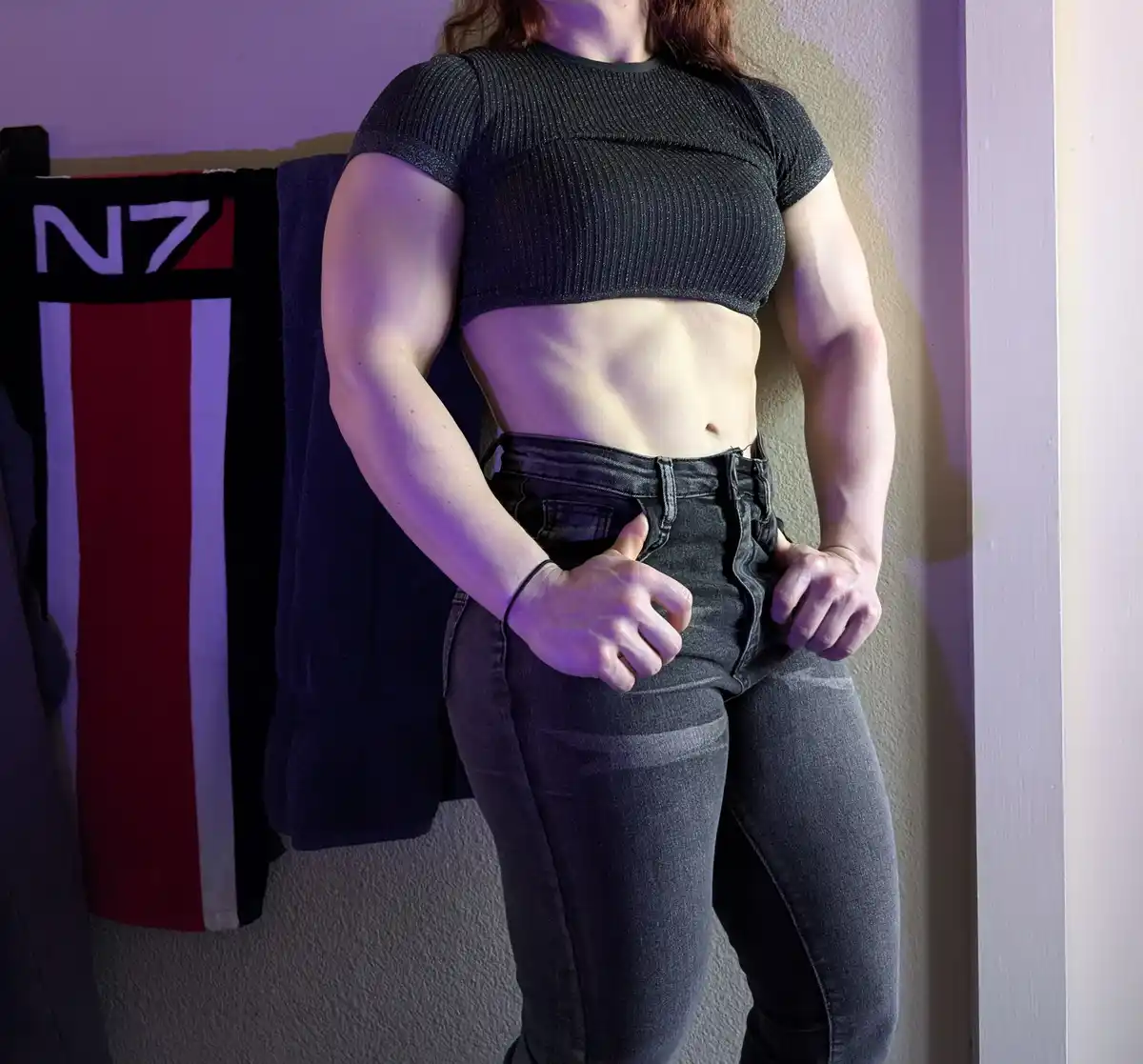 A person with a muscular build, wearing a black crop top and dark jeans, stands with hands on hips, showcasing defined arms…