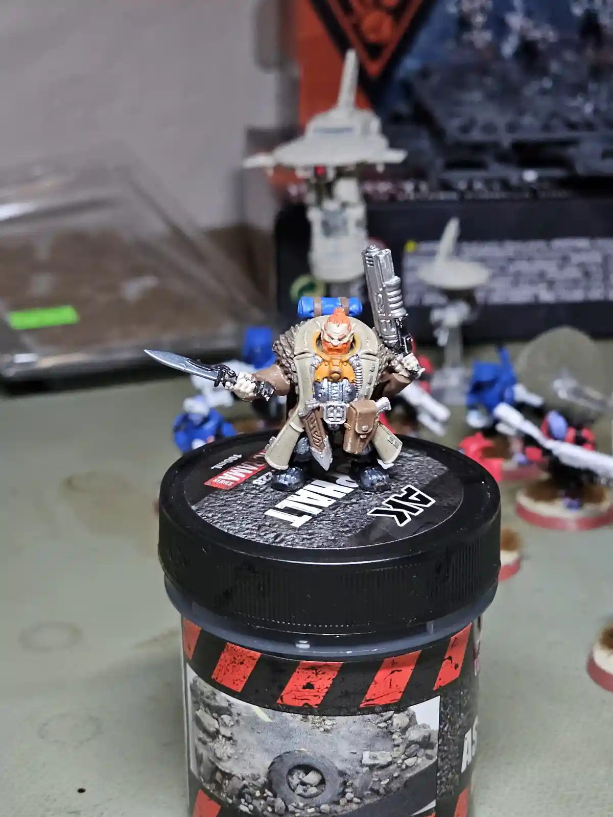 A painted miniature figurine of a fantasy warrior with orange hair, wielding a sword and a pistol, stands atop the black lid…