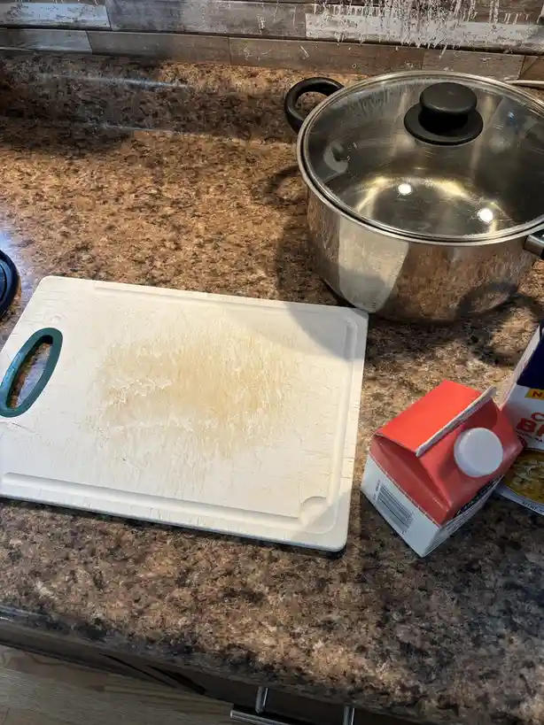 A heavily scratched and stained white plastic cutting board with a green handle sits on a brown granite kitchen counter next…