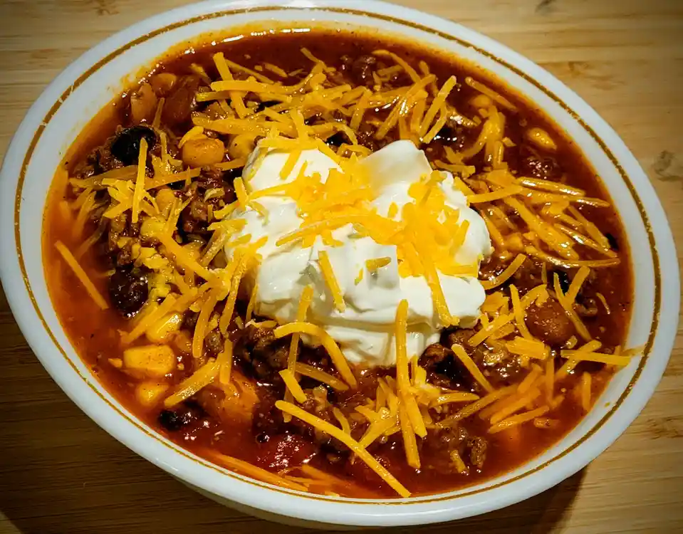 A close-up photo of a white bowl filled with chili, topped with shredded cheddar cheese and a dollop of sour cream, sitting…