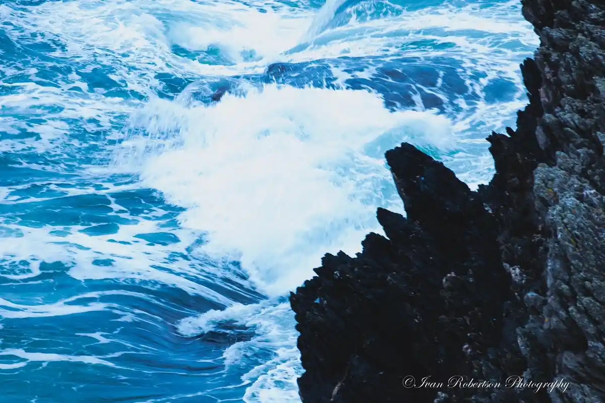 A dynamic photograph shows powerful blue ocean waves crashing against dark, jagged rocks on the right side of the frame…
