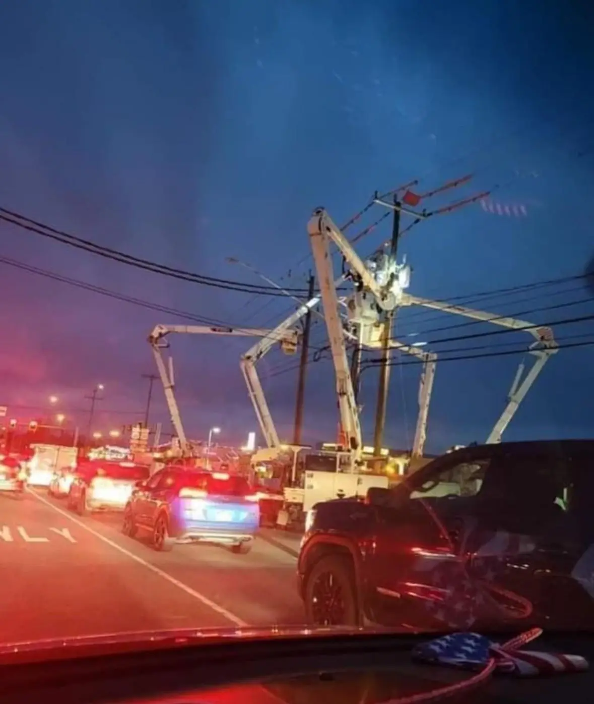 a nighttime photo taken from inside a car shows multiple utility trucks with extended booms surrounding a utility pole…