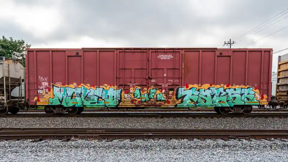A red freight train car covered in vibrant green and yellow graffiti sits on railway tracks under a cloudy sky. The overlaid…
