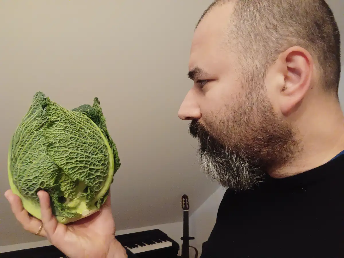 A man with a beard and short hair holds a large green savoy cabbage in his left hand, looking at it with a skeptical or…