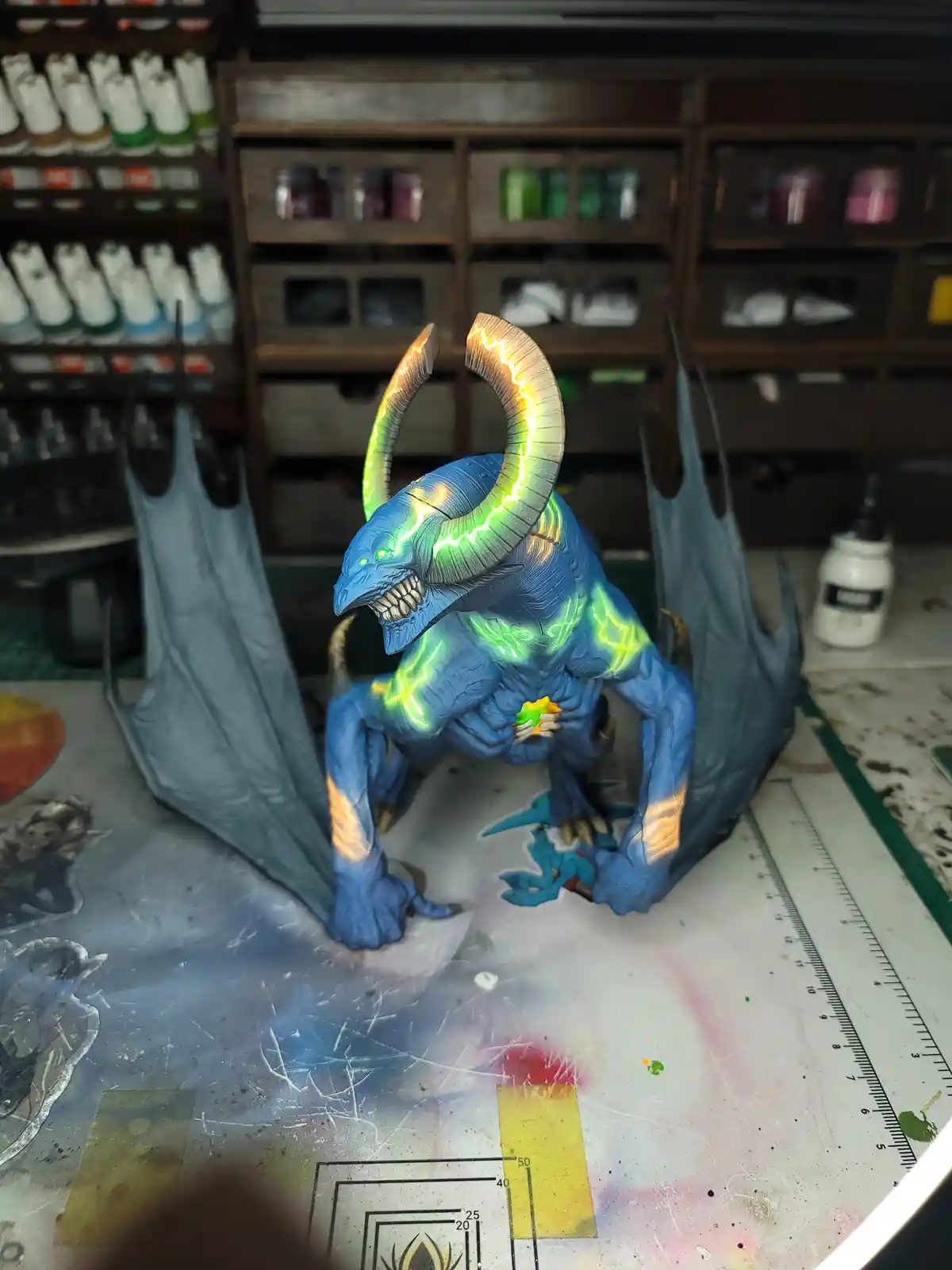 A blue, horned, demonic figurine with glowing green lightning patterns on its body and wings is posed on a paint-splattered…