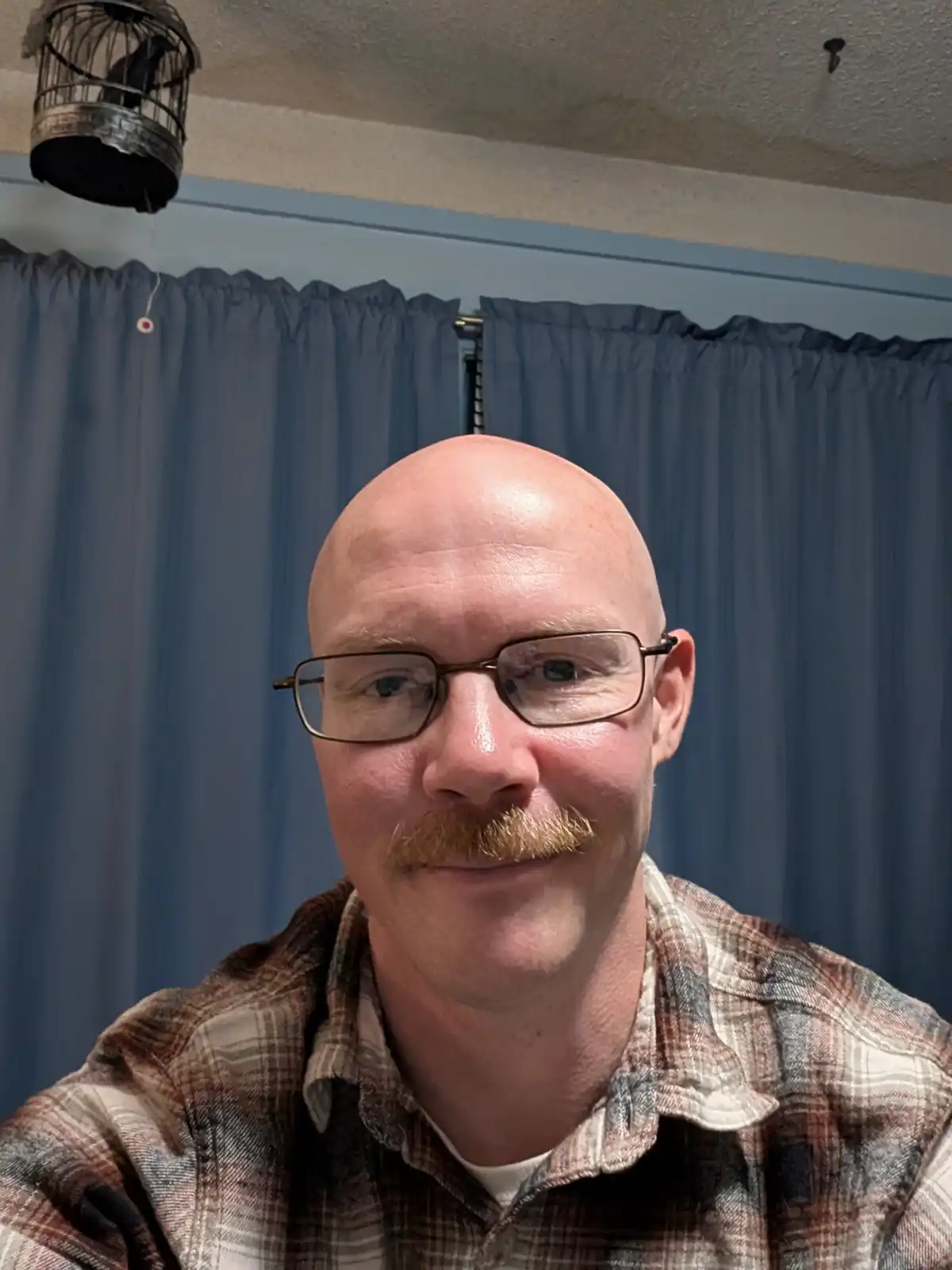 A bald man with glasses and a mustache smiles at the camera, wearing a plaid shirt. Behind him are blue curtains and a…