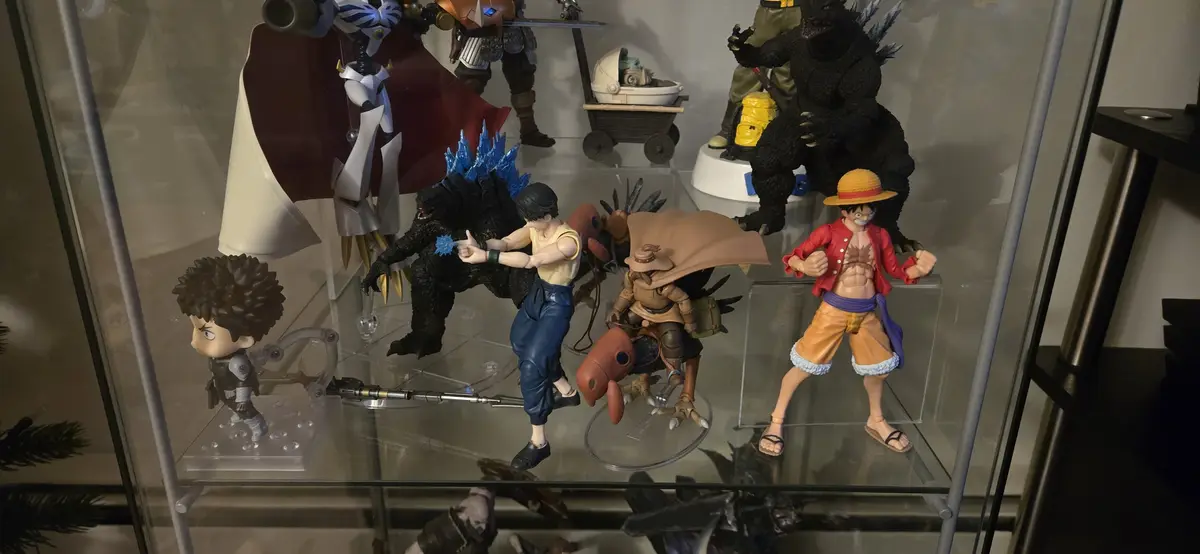A glass display case holds various anime and movie action figures, including Godzilla, Luffy, and other characters, with a…