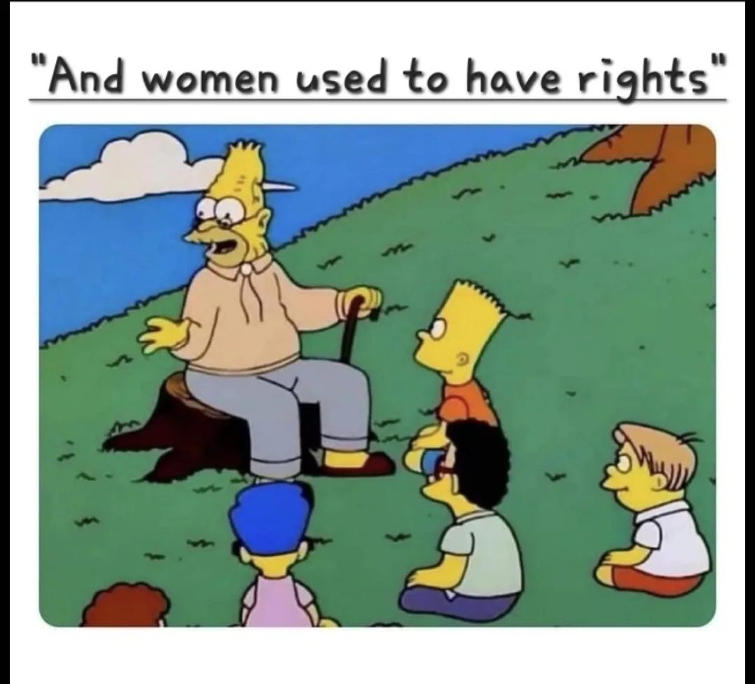 An older, balding yellow-skinned man with a cane sits on a tree stump outdoors, gesturing while speaking to a group of…