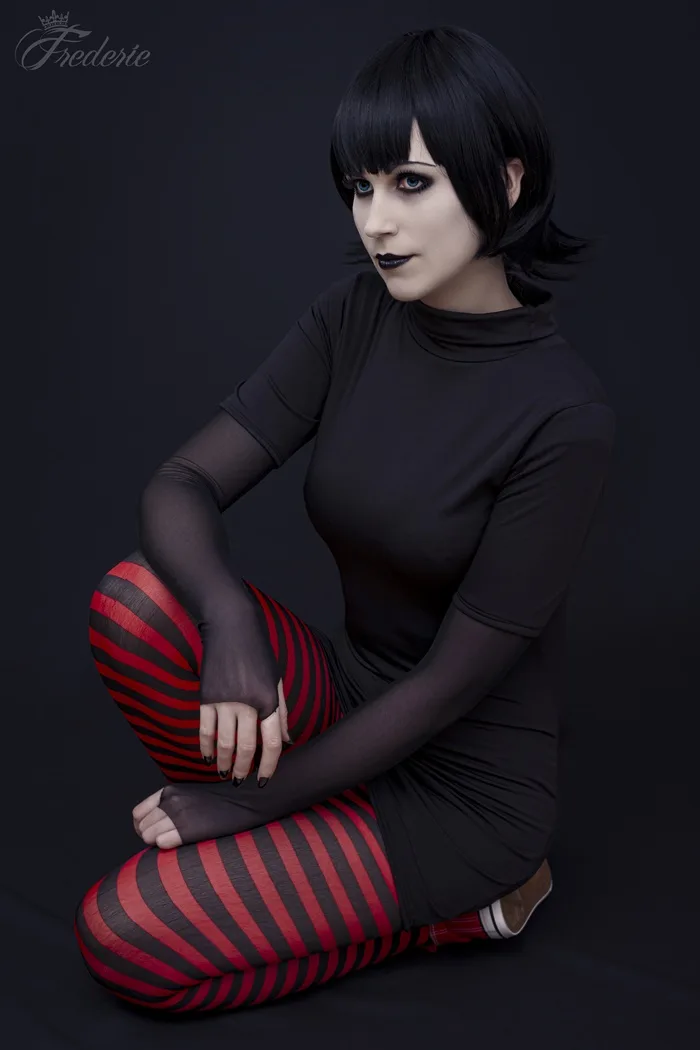 A person with short black hair and heavy black makeup sits cross-legged on a black background, wearing a black turtleneck…