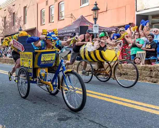 Two people in a parade ride decorated bicycles, one with a 'Sheppard Realty' sign and the other with a 'Meader' barrel, both…