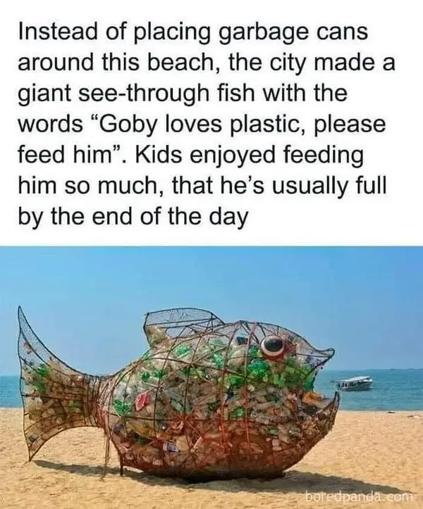 A large, transparent fish sculpture made of plastic bottles and wire sits on a sandy beach near the ocean. The fish has a…