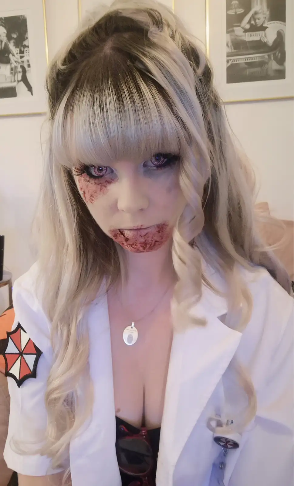 A person with long blonde hair and dramatic makeup, including red blood-like stains around their eyes and mouth, wears a…