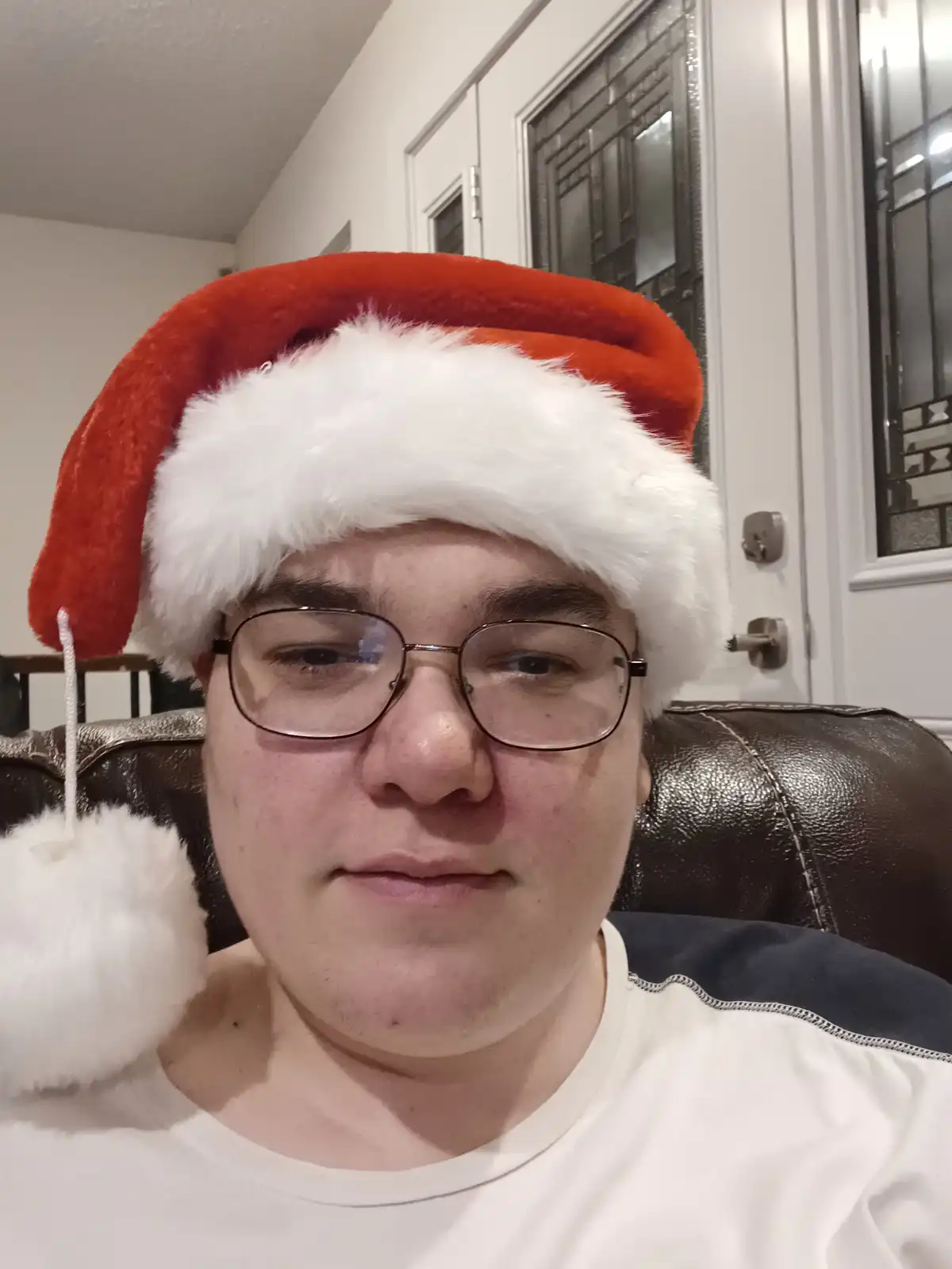 A person wearing a red and white Santa hat and glasses sits in a brown leather chair indoors, looking directly at the camera…